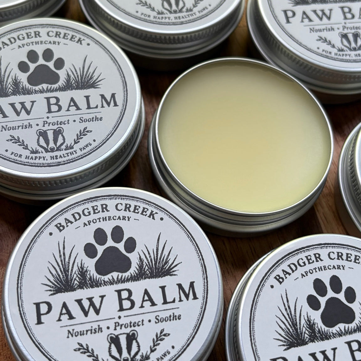 Several round silver tins of Paw Balm by Badger Creek Apothecary are arranged on a wooden surface, with one tin open to show a pale yellow balm inside.