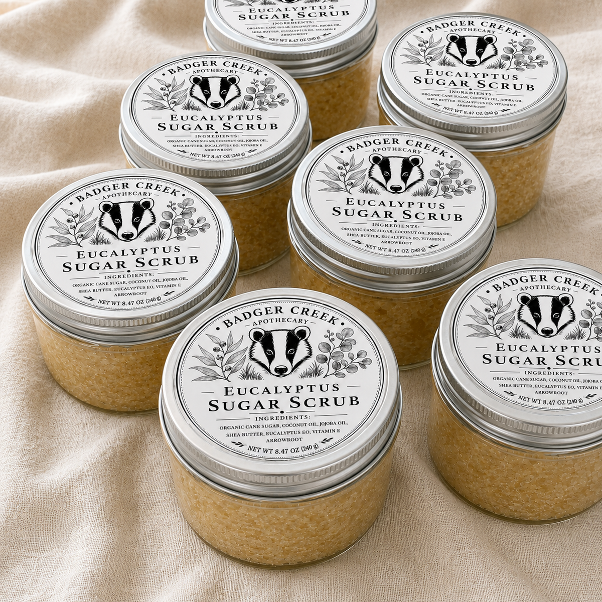 Six jars of Badger Creek Apothecary Eucalyptus Sugar Scrub arranged on a beige fabric background. Each clear jar contains a light tan scrub and is topped with a silver metal lid featuring a white label with a black badger illustration and product name.