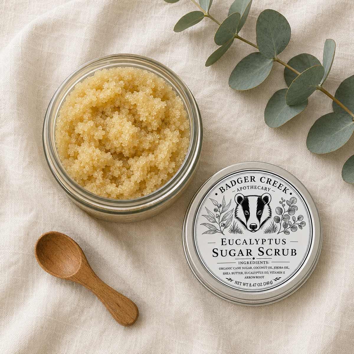 An open glass jar filled with coarse pale yellow sugar scrub sits beside its labeled lid on a cream linen surface, with a small wooden spoon and eucalyptus leaves around it. The lid reads Badger Creek Apothecary and Eucalyptus Sugar Scrub.
