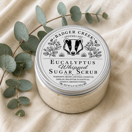 A round jar of Badger Creek Apothecary Eucalyptus Whipped Sugar Scrub sits on a beige fabric surface with eucalyptus leaves around it. The lid has a white label with black text and a badger illustration.