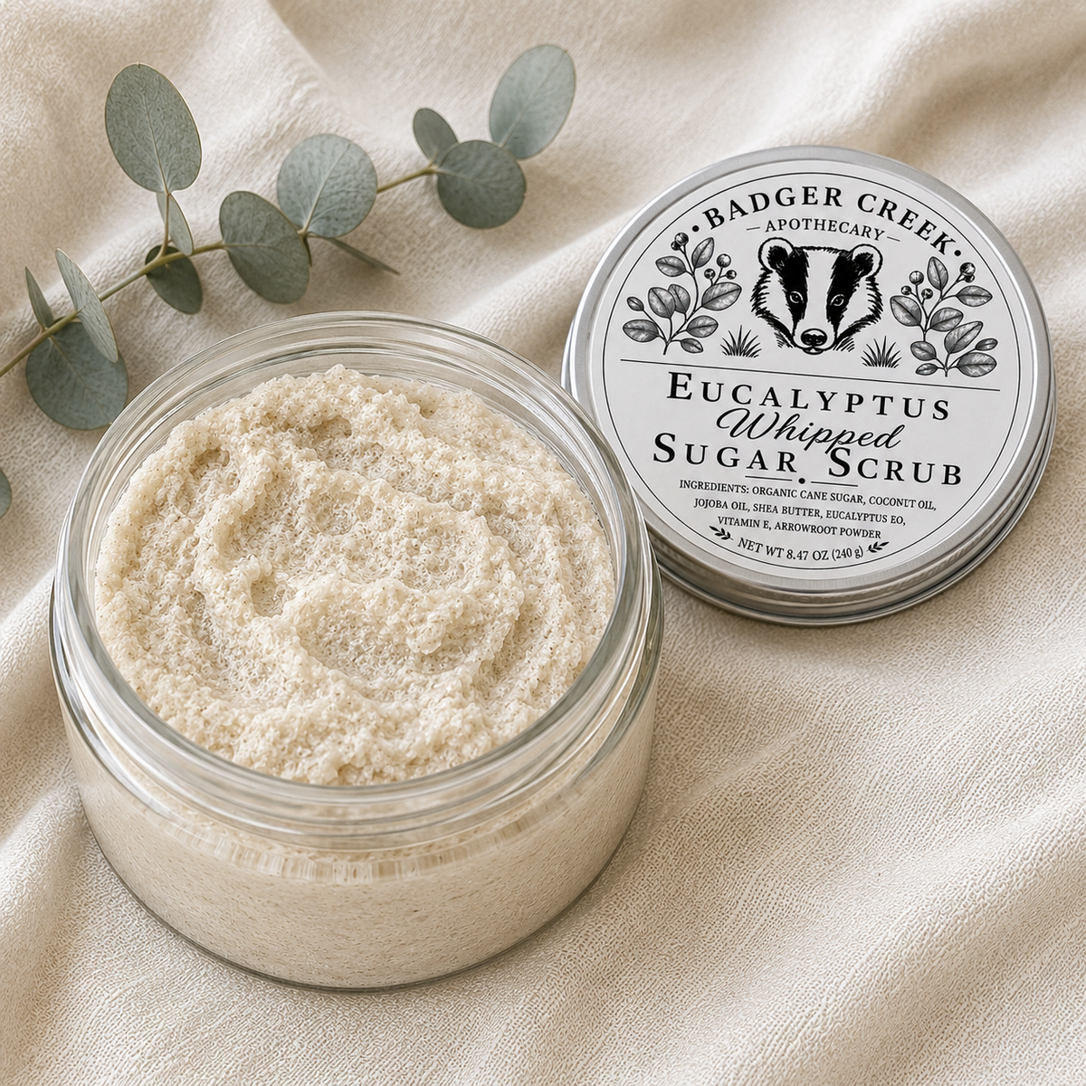 An open glass jar of Badger Creek Apothecary Eucalyptus Whipped Sugar Scrub sits on a beige fabric surface next to its silver and white metal lid. A sprig of eucalyptus leaves is placed behind the jar.