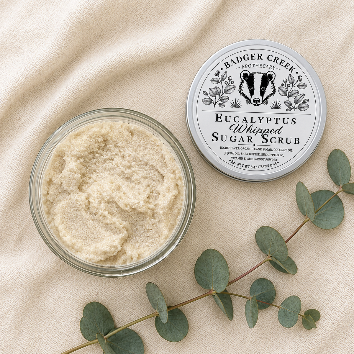 An open round glass jar of Badger Creek Apothecary Eucalyptus Whipped Sugar Scrub sits beside its silver lid on a beige fabric surface, with eucalyptus leaves arranged around them.