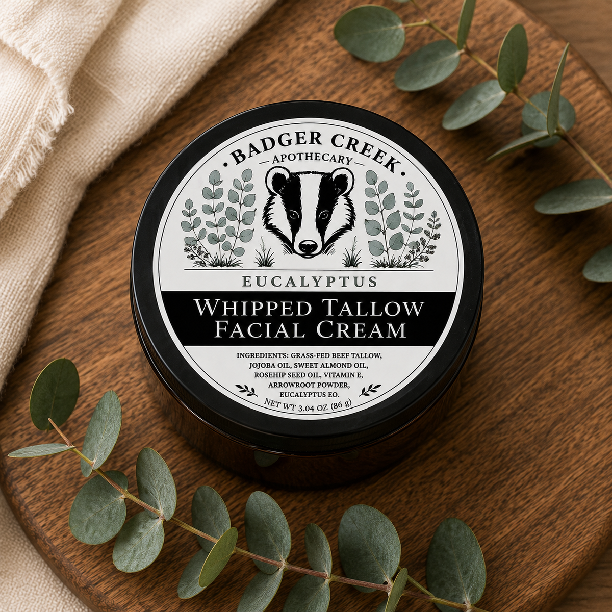 A round black tin of Badger Creek Apothecary Whipped Tallow Facial Cream sits on a round wooden board, surrounded by eucalyptus leaves and a beige fabric napkin.