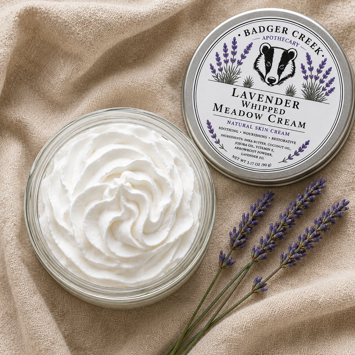 An open round glass jar filled with whipped cream sits beside the product lid on a beige fabric surface, with lavender sprigs placed in front. The lid shows the Badger Creek Apothecary branding and reads Lavender Whipped Meadow Cream.