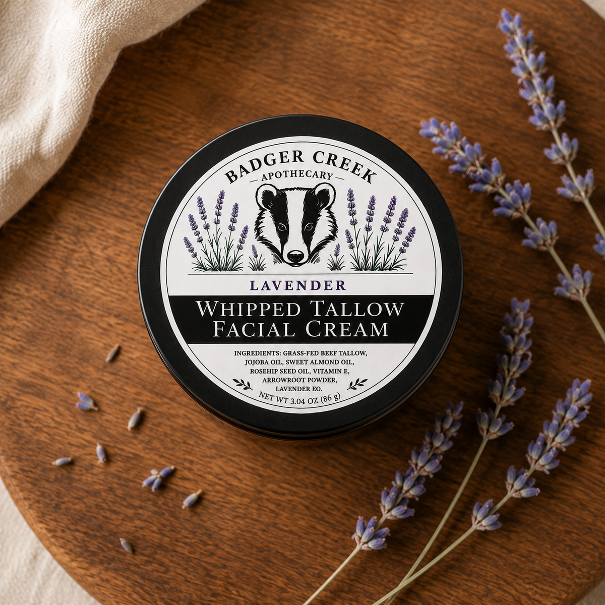 A round container of Badger Creek Apothecary Whipped Tallow Facial Cream sits on a wooden surface, surrounded by lavender sprigs and a folded cream fabric. The front label features a black-and-white badger illustration with lavender flowers and the text 'Lavender'.