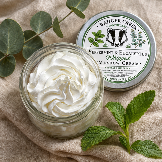 An open glass jar filled with whipped cream sits on a beige fabric surface beside the product lid, with eucalyptus leaves and mint sprigs around it. The lid reads Badger Creek Apothecary and Peppermint & Eucalyptus Whipped Meadow Cream.