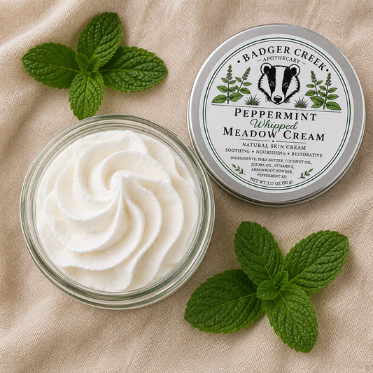 An open glass jar filled with whipped white cream sits beside the product lid on a beige fabric background, surrounded by fresh mint leaves. The lid label reads Badger Creek Apothecary Peppermint Whipped Meadow Cream.