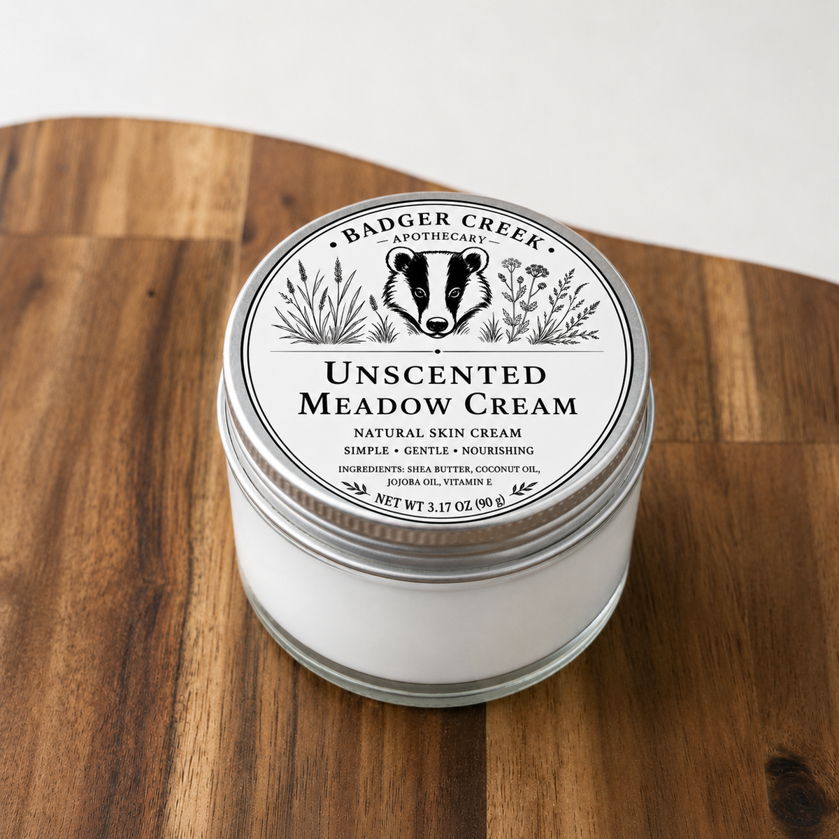 A glass jar of Badger Creek Apothecary Unscented Meadow Cream sits on a wooden surface. The jar is filled with white cream and has a silver metal lid with a white paper label featuring a black badger illustration.