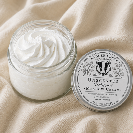 An open glass jar filled with whipped cream sits on beige fabric next to its silver lid, which features a black-and-white badger illustration and the text 'Badger Creek Apothecary Unscented Whipped Meadow Cream'.