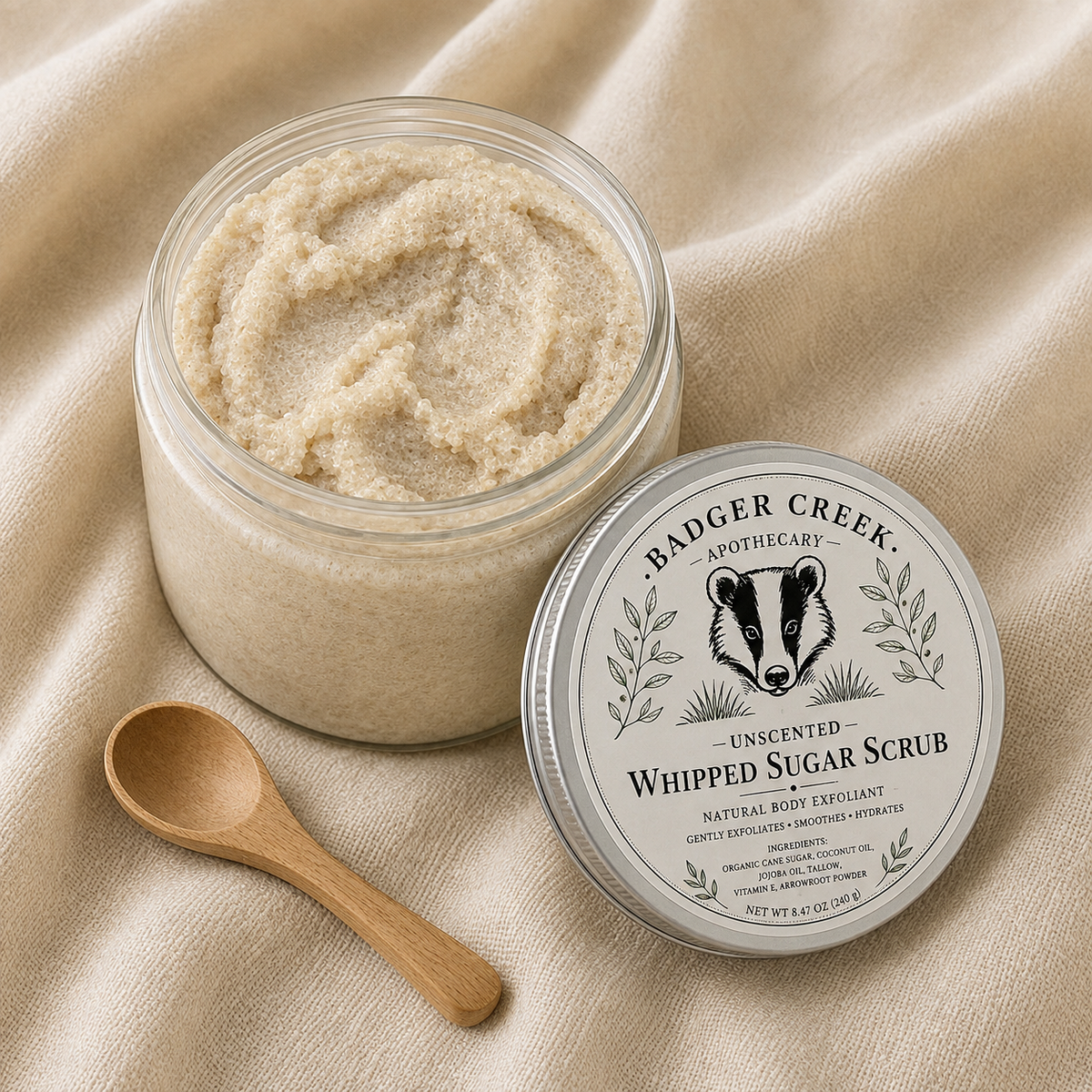 An open glass jar of Badger Creek Apothecary Whipped Sugar Scrub sits on draped beige fabric next to its silver lid and a small wooden spoon.