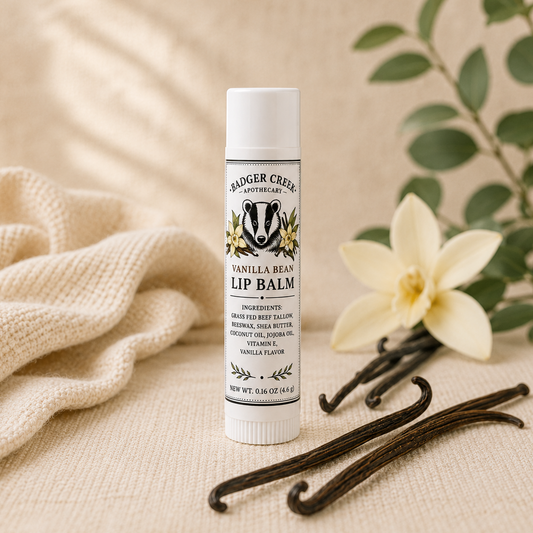 A tube of Vanilla Bean Lip Balm stands upright on a beige fabric surface, styled with a cream knit cloth, a white flower, green leaves, and vanilla beans.